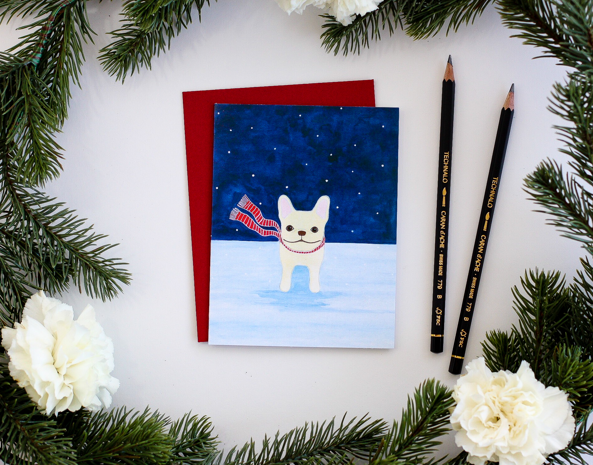 A flat-lay of a holiday card surrounded by pine needles and pencils