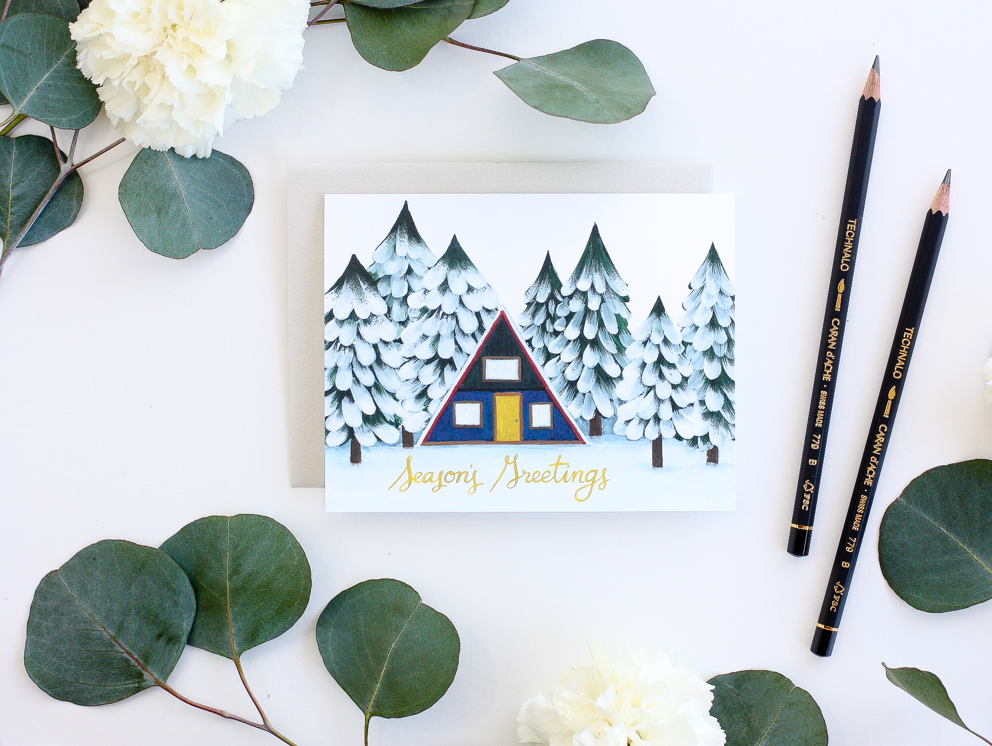 Flat-lay of an illustrated holiday card surrounded by eucalyptus leaves and pencils